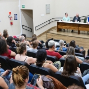 Residentes sentados no auditório observam a mesa de abertura do evento. Secretária Ana Costa está ao microfone. 