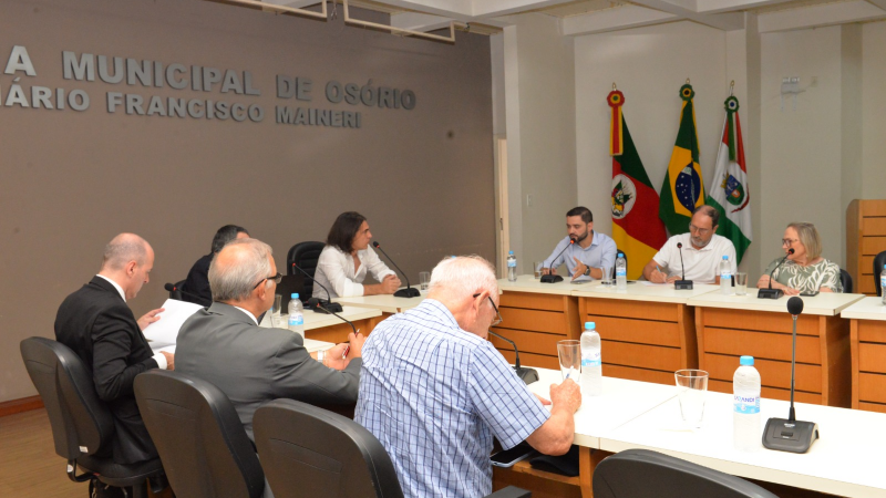 Autoridades na câmara sentado à mesa. A mesa é branca com base em madeira. A parede ao fundo é cinza, com letreiro escrito "Câmara Municipal de Osório". As bandeiras do estado do Rio Grande do Sul, do Brasil e do município de Osório também aparecem no centro da imagem.