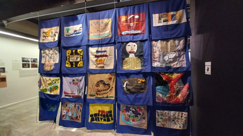 As peças de tapeçaria tem fundo azul e são bordadas cada uma com um desenho diferente. Estão agrupadas em cinco fileiras, suspensas como num varal. Ao fundo o ambiente de uma galeria de arte, com piso e paredes brancas.