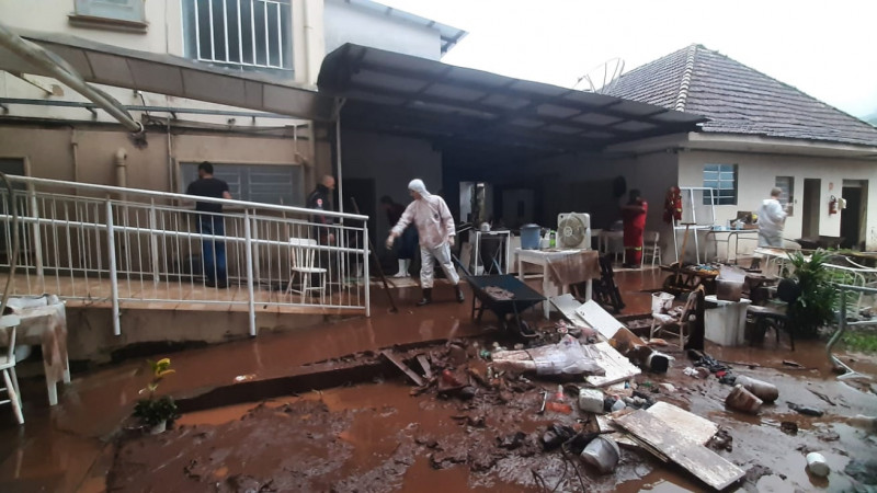Fundos do hospital completamente cheios de lama, logo após baixar a água da enchente. Algumas pessoas estão numa rampa de acesso iniciando trabalhos de limpeza. Muitos objetos e materiais estão ao chão, na lama. O dia aparece nublado.