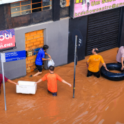 Área alagada da cidade, em uma esquina com prédios de comércio e residenciais. Homens carregam sacolas, uma caixa de isopor e uma boia na inundação.