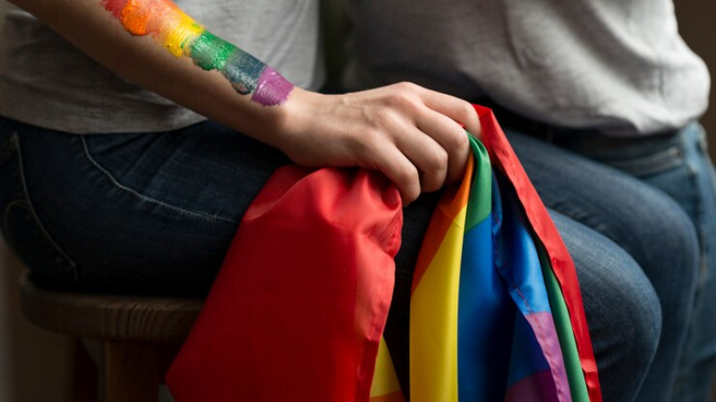 Imagem de uma pessoa, apenas da cintura para baixo, sentada, vestindo uma camiseta cinza e uma calça jeans. Ela segura uma bandeira do orgulho LGBT+ e tem o braço pintado nas mesmas cores do arco íris.
