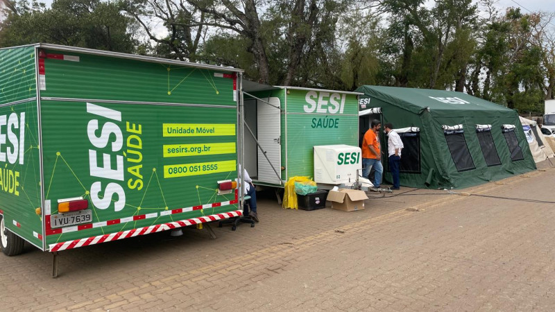 Local onde a tenda foi instalada com unidade móvel do Sesi estacionada ao lado. Todas as estruturas são na cor verde escuro e trazem os dizeres em caixa alta "SESI SAÚDE". Ao fundo estão algumas árvores.