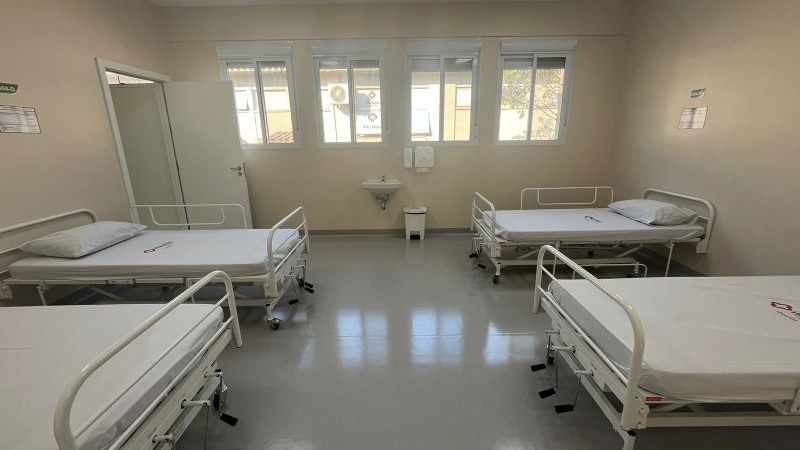 Quatro camas hospitalares, duas do lado esquerdo e duas do lado direito. Ao fundo, janelas abertas