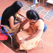 Duas mulheres indígenas estão sentadas em cadeiras escolares na cor azul. A mulher à esquerda, de cabelos lisos, longos e soltos desenha o braço a outra mulher, que tem os cabelos trançados. Ao fundo está um chão de quadra de escola, em tom laranja. Um desenho de números no chão forma a "amarelinha". 