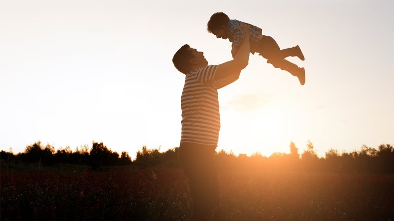 Um homem (pai) erguendo uma criança (filho) com o pôr do sol e árvores ao fundo