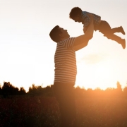 Um homem (pai) erguendo uma criança (filho) com o pôr do sol e árvores ao fundo