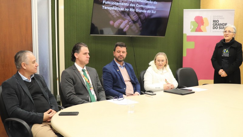 Da esquerda para a direita, estão sentados à mesa Roque Serpa, Fabrício Peruchin, Neri, O Carteiro e Arita Bergmann. Na ponta da mesa, vestindo uma roupa preta, está Rosângela Scurssel. A mesa é bege clara. Ao fundo tem uma parede verde e uma tela onde está o início da apresentação da nova regulamentação. No canto direito está o banner do governo do estado.