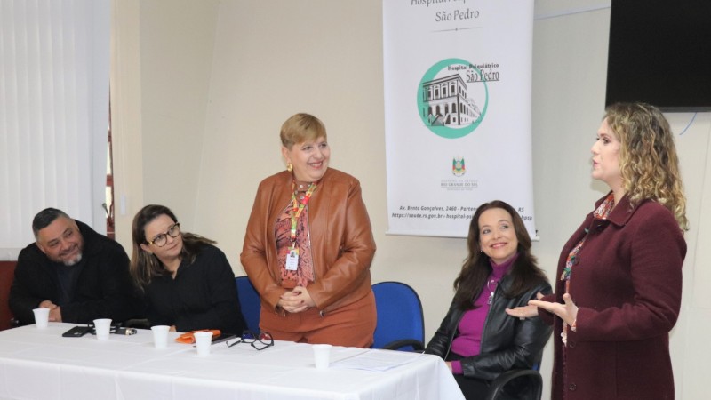 Um homem e quatro mulheres, duas em pé e duas sentadas em frente a uma mesa
com toalha branca e copos de plástico branco em cima da mesa, um banner atrás com ao fundo branco e com a figura do hospital São Pedro em Preto e letras pretas.
