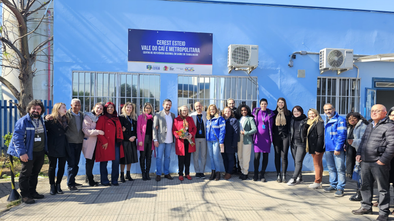 Equipe do Centro de Referência e autoridades presentes no evento posam para foto em frente ao prédio do serviço. O prédio é azul claro e tem uma placa padronizada em azul escuro com os dizeres em letras brancas maiúsculas: CEREST ESTEIO VALOR DO CAÍ E METROPOLITANA. O dia está ensolarado e tem sombra das árvores sobre as pessoas na foto.