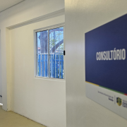 Porta de um consultório mostra um ambiente todo branco, com duas janelas no ambiente. Na porta uma placa padronizada em azul escuro tem escrito em letras maiúsculas brancas: CONSULTÓRIO 2.