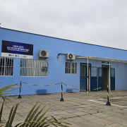 Prédio do centro todo pintado em azul claro, com portas em tom de azul escruto e três janelas com grades brancas. O chão é de pedras cinzas. O dia está nublado e uma folhagem está em primeiro plano na imagem. 