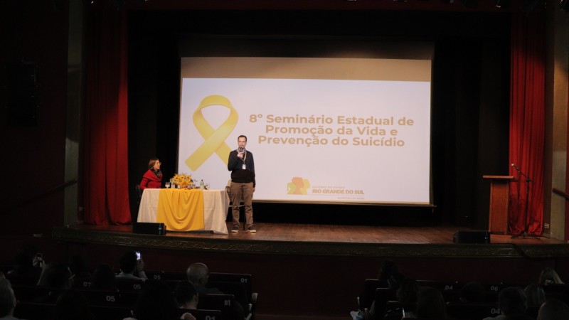 Um palco de teatro com fundo escuro e cortinas vermelhas nos cantos tem uma tela de projeção bem ao centro. Nela, está projetado em fundo branco e com as letras em cor preta: 8º Seminário Estadual de Promoção da Vida e Prevenção do Suicídio", com o logo do governo do Rio Grande do Sul abaixo. Um laço amarelo está à esquerda da imagem projetada. À frente, com microfone, um homem branco está falando. Ele veste uma calça bege e um suéter azul marinho. Uma mesa com toalha branca e uma toalha amarela por cima está no canto esquerdo da imagem. Sentada à esta mesa está uma mulher branca, de cabelos loiros na altura dos ombros, vestindo um casaco vermelho.