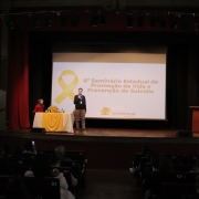 Um palco de teatro com fundo escuro e cortinas vermelhas nos cantos tem uma tela de projeção bem ao centro. Nela, está projetado em fundo branco e com as letras em cor preta: 8º Seminário Estadual de Promoção da Vida e Prevenção do Suicídio", com o logo do governo do Rio Grande do Sul abaixo. Um laço amarelo está à esquerda da imagem projetada. À frente, com microfone, um homem branco está falando. Ele veste uma calça bege e um suéter azul marinho. Uma mesa com toalha branca e uma toalha amarela por cima está no canto esquerdo da imagem. Sentada à esta mesa está uma mulher branca, de cabelos loiros na altura dos ombros, vestindo um casaco vermelho.