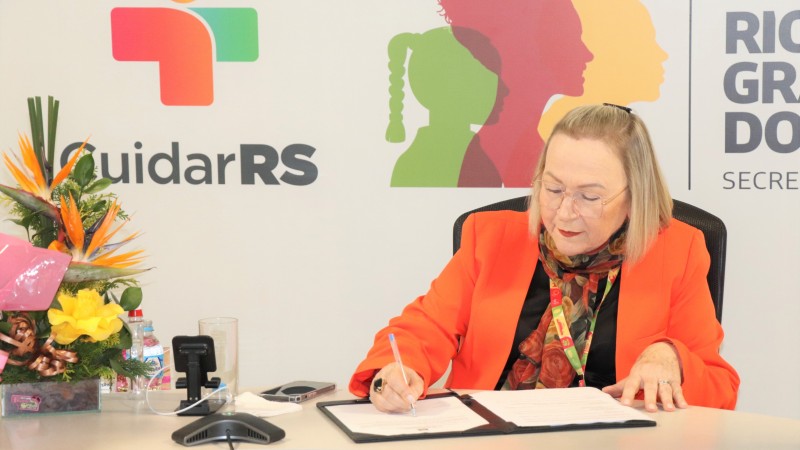 Arita Bergmann assina o convênio sobre uma mesa bege. Ela é uma mulher branca, com cabelos loiros na altura dos ombros e usa um óculos de grau de armação transparente. Está vestindo um blazer laranja e um lenço florido em tons alaranjados no pescoço. Ao fundo está um painel com o slogan de governo. Um arranjo de flores está no canto esquerdo da imagem.