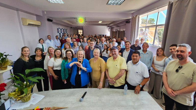 Atrás de uma mesa retangular cerca de 100 pessoas em pé olhando e sorrindo. A secretária Arita Bergmam está junto com as pessoas, posicionada na fila da frente. na frente 