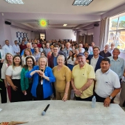 Atrás de uma mesa retangular cerca de 100 pessoas em pé olhando e sorrindo. A secretária Arita Bergmam está junto com as pessoas, posicionada na fila da frente. na frente 