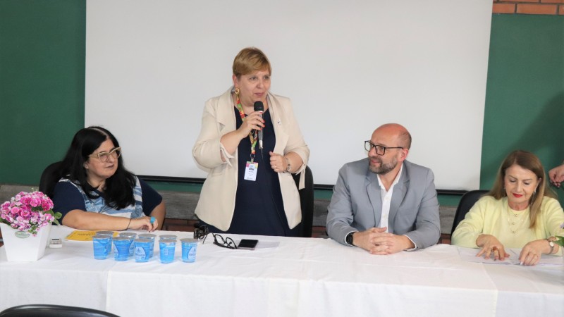 Secretária Ana Costa ao centro segura o microfone. Ela é uma mulher branca, de cabelos loiros e curtos. Veste uma blusa preta e um blazer bege. No lado direito uma mulher branca de cabelos pretos está sentada. No lado esquerdo estão um homem branco, de terno cinza e uma mulher branca, loira com cabelo na altura dos ombros, vestindo uma blusa amarela. Todos estão atrás de uma mesa com uma toalha branca. Ao fundo um quadro de sala de aula e uma tela de projeção.