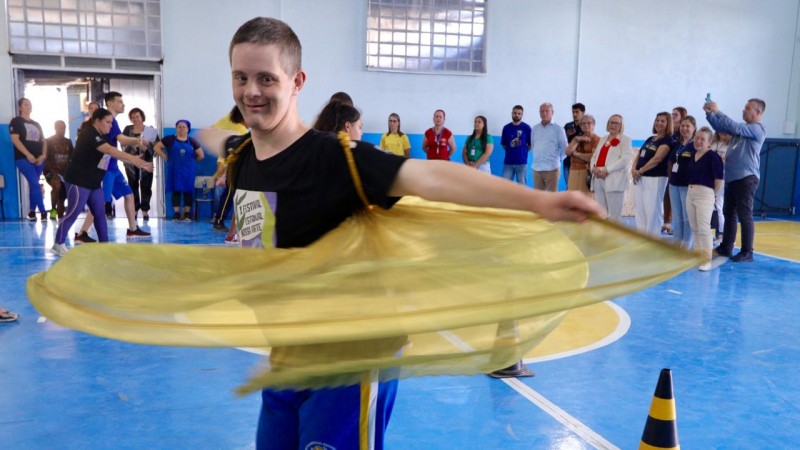 Um menino dançando com um adereço amarelo que gira o tecido em forma de bambolê ele está em um ginásio e tem várias pessoas ao fundo.