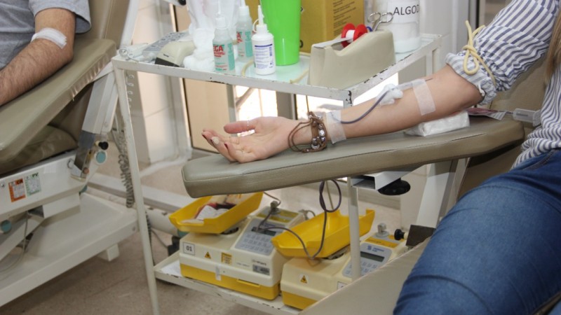 Pessoa sentada na cadeira do hemocentro com o braço com aparelho de coleta em local com equipamentos de doação de sangue. 