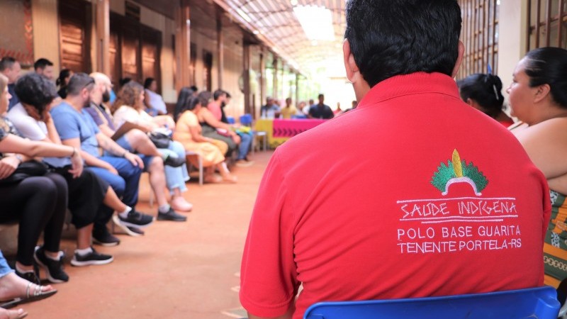 Um homem virado de costas está olhando para a frente. Ele veste uma camiseta vermelha com os dizeres em letras maiúsculas, na cor branca: saúde indígena - polo base Guarita Tenente Portela-RS. Outras pessoas aparecem desfocadas na imagem.