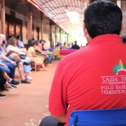 Um homem virado de costas está olhando para a frente. Ele veste uma camiseta vermelha com os dizeres em letras maiúsculas, na cor branca: saúde indígena - polo base Guarita Tenente Portela-RS. Outras pessoas aparecem desfocadas na imagem.