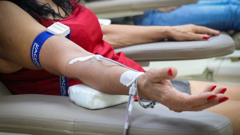 Um braço de mulher que está sentada com blusa vermelha e com aparelho nas veias fazendo coleta de sangue