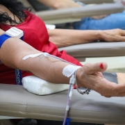 Um braço de mulher que está sentada com blusa vermelha e com aparelho nas veias fazendo coleta de sangue