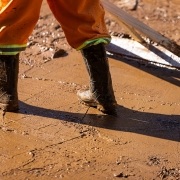 Imagem mostra parte inferior da pernas de uma pessoa, com calças e botas de equipamentos de proteção individual. A pessa empurra uma lama com uma ferramenta de madeira 
