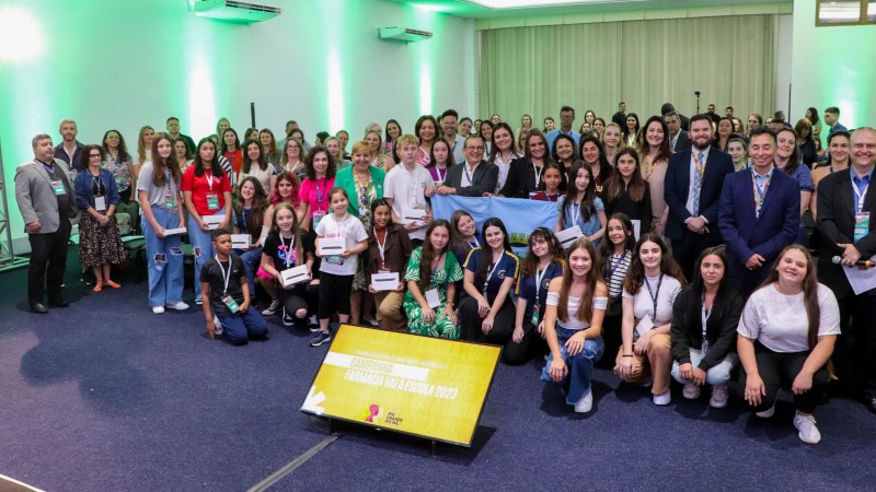 Nu auditório iluminado com luzes verdes e chão de carpete cinza, crianças e adolescentes e seus professores posam para foto com autoridades durante a premiação.