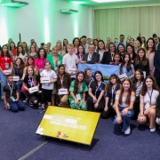 Nu auditório iluminado com luzes verdes e chão de carpete cinza, crianças e adolescentes e seus professores posam para foto com autoridades durante a premiação.