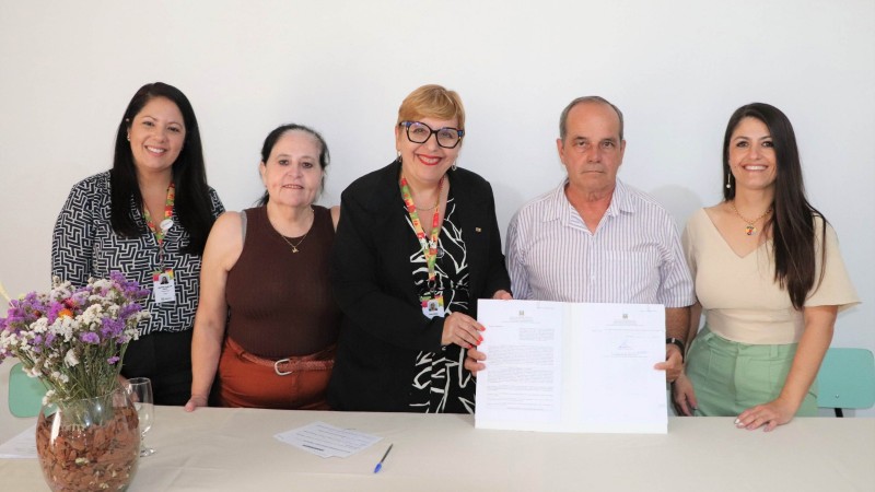 Cinco pessoas ( quatro mulheres e um homem), mostrando o documento assinado em frente a uma mesa com um vaso de flores brancas e lilás em cima, ao lado. 