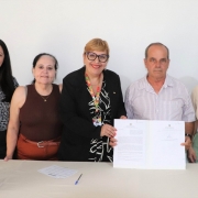 Cinco pessoas ( quatro mulheres e um homem), mostrando o documento assinado em frente a uma mesa com um vaso de flores brancas e lilás em cima, ao lado. 