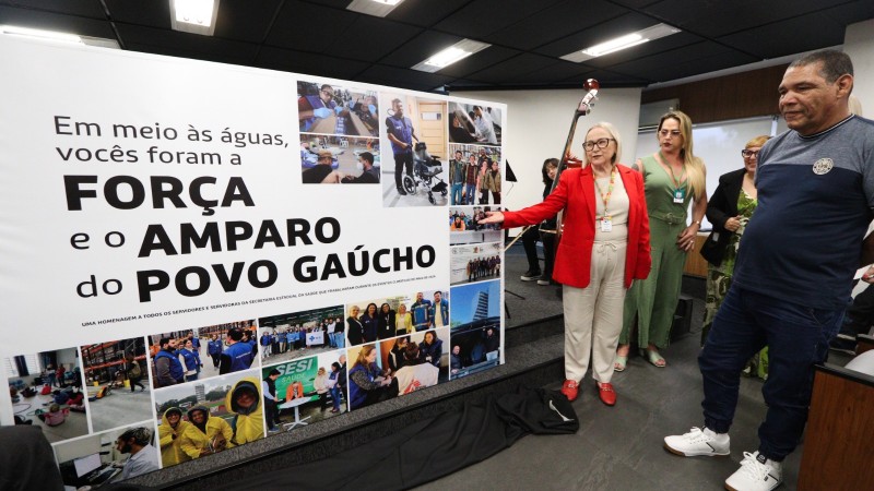 Três mulheres e um homem em pé em frente ao painelcom fotos em homenagem aos servidores pela atuação nas enchentes, com a frase Em meio às [aguas, vocês foram a força e o amparo do povo gaúcho