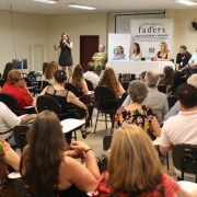 Uma mulher em cima do palco, com vestido preto, falando ao microfone ao lado de uma mesa com toalha branca, inde seis  pessoas estão sentadas, quatro mulheres e dois homens. Em frente, aparece um público formado por cerca de vintes pessoas. 