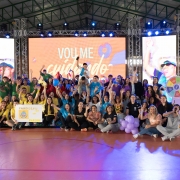 Adolescentes participantes da última edição do Geração Consciente posam para foto em um ginásio. Eles vestem roupas coloridas em tons de azul, amarelo e verde, representando as equipes. Alguns seguram cartazes com os nomes das escolas. Ao fundo um painel dividido em três telas exibe a projeção com frases e imagens do programa.