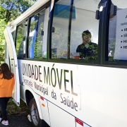 Unidade móvel da Secretaria Municipal de Saúde de Osório está estacionada. Ao fundo da foto um homem vestindo calça jeans e camiseta preta conversa com uma mulher de colete laranja. A unidade móvel é um ônibus branco e tem uma folha de ofício em uma janela indicando os testes rápidos que são realizados: HIV, sífilis, hepatite B e hepatite C.