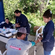 Uma mulher de jaleco azul faz aferição de pressão arterial em um homem de camiseta cinza e boné cinza. Ao fundo a mesa de enfermagem e outros profissionais de saúde realizam procedimentos.