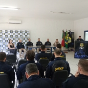 Uma sala de fundo branco e mesa de vidro ao centro. Está composta uma mesa de autoridades da Polícia Rodoviária Federal e Secretaria da Saúde. Policiais rodoviários estão sentados de frente para a mesa. Um cerimonialista da PRF conduz a fala no púlpito no canto direito.