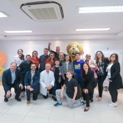 Ao centro da foto a secretária Arita está acompanhada de diretores, equipe do ICI e crianças pacientes do instituto. O médico Algemir Brunetto, o cantor Rafael Malenotti e o leão, mascote do ICI, também estão posando junto do grupo. O ambiente ao fundo é todo na cor branca.