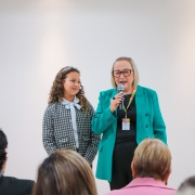 Secretária Arita fala ao microfone. Ela é uma mulher branca, de cabelos loiros na altura dos ombros, usa óculos de grau de armação escura, veste uma roupa preta e um blazer na cor verde. À sua esquerda estás Helena, uma menina branca de cabelos castanhos e cacheados. Ela veste uma roupa quadriculada na cor cinza e olha para Arita com sorriso tímido. Ao fundo uma parede branca.