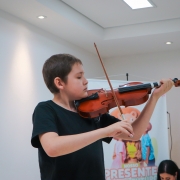 Gabriel toca violino. Ele é um menino branco, de cabelos castanhos claros e curtos. Veste uma camiseta na cor preta.