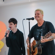 Arthur está segurando o microfone e cantando. Ele é um menino branco, de cabelos pretos e curtos. Ao seu lado está o cantor Rafael Malenotti tocando violão. Ele é um homem branco, de cabelos curtos e loiros. O fundo é uma parede na cor branca.
