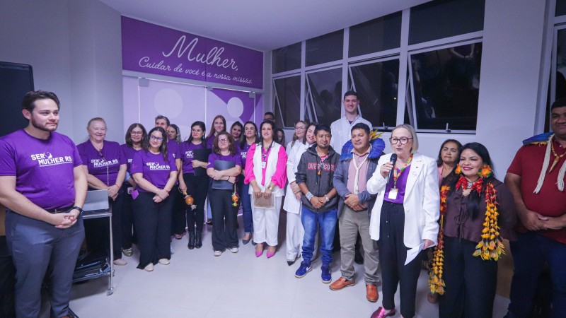 Um grupo de pessoas com camisas lilás e outras com adereços indígena com a secretária Arita Bergmann discursando na recepção do serviço SER Mulher