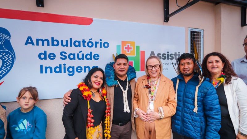 A secretária Arita está ao centro da foto, sorrindo. Ela é uma mulher branca, de cabelos loiros na altura dos ombros. Usa um conjunto na cor caqui. Está acompanhada de lideranças indígenas com vestes características. Todos sorriem para a câmera. Ao fundo uma placa na cor branca, com o logo do Programa Assistir e os dizeres em letra na cor azul: Ambulatório de Saúde Indígena. Uma parede salmão está ao fundo.