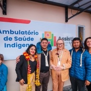 A secretária Arita está ao centro da foto, sorrindo. Ela é uma mulher branca, de cabelos loiros na altura dos ombros. Usa um conjunto na cor caqui. Está acompanhada de lideranças indígenas com vestes características. Todos sorriem para a câmera. Ao fundo uma placa na cor branca, com o logo do Programa Assistir e os dizeres em letra na cor azul: Ambulatório de Saúde Indígena. Uma parede salmão está ao fundo.