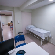 Uma sala de atendimento contém uma mesa branca, uma cadeira azul e uma maca. A porta da sala está aberta.
