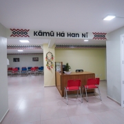 O hall de entrada do ambulatório é em tons claros. Traz uma mensagem escrita em uma língua originária. Uma mesa de madeira e cadeiras vermelhas estão no ambiente.