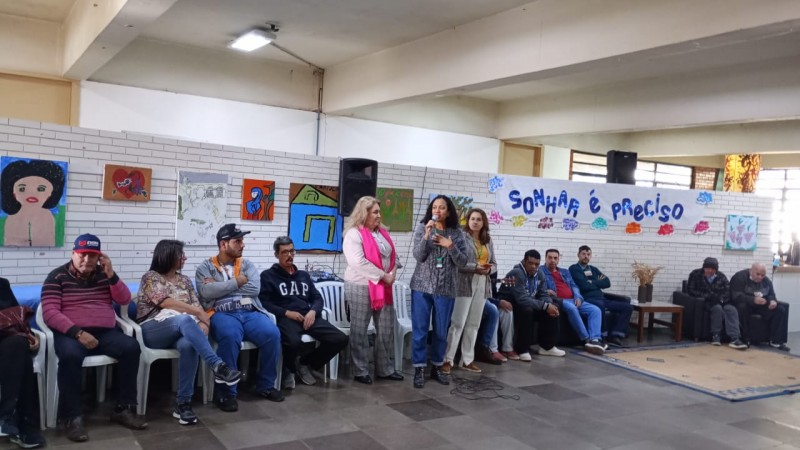 Uma mulher negra fala ao microfone. Ela tem cabelos escuros e cacheados na altura dos ombros, veste um casaco cinza e uma calça jeans azul. Está acompanhada de outras duas mulheres, brancas, que ouvem ela falar. O ambiente tem cadeiras e pessoas sentadas. Um muro de tijolos brancos está trás com alguns cartazes e quadros expostos. Uma faixa com a frase "sonhar é preciso" em letras azuis também compõe o cenário. O chão é de pedra na cor cinza.