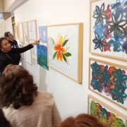 Uma mulher negra, vestindo uma roupa preta, aponta para um quadro na parede. A parede branca tem várias obras expostas e pessoas observando.